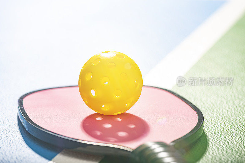 A pink Pickleball paddle with a yellow ball sitting on the court – lens flare